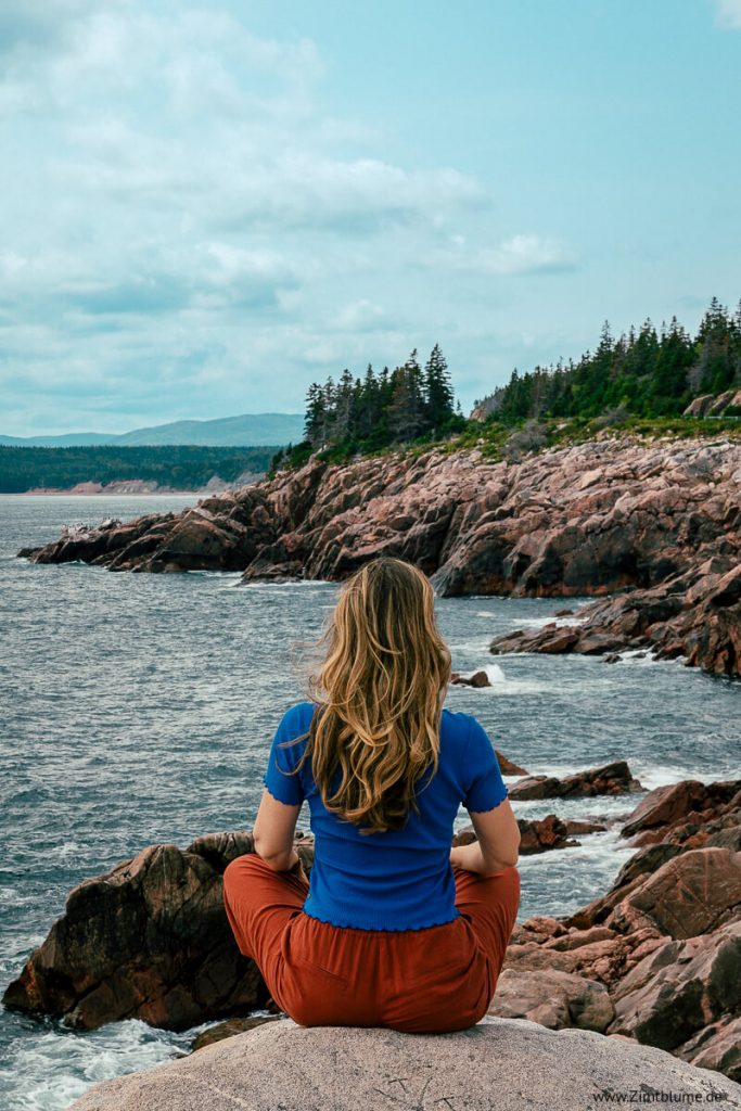 Angelina von Zimtblume sitzt auf einem Felsen bei Green Cove am Cabot Trail und blickt auf das Meer und die Klippen.