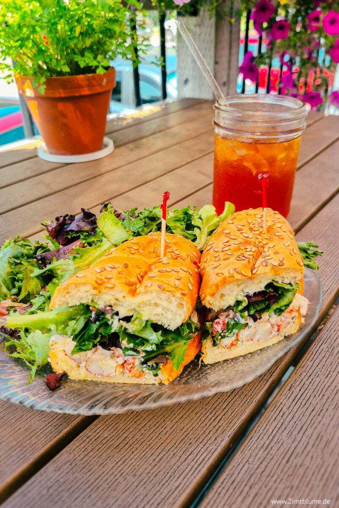 Aufgeschnittene Lobster Roll und ein Glas Eistee auf einem Holztisch im Außenbereich des Kiwi Cafe in Chester, Nova Scotia, umgeben von Petunien und Kräutertöpfen.